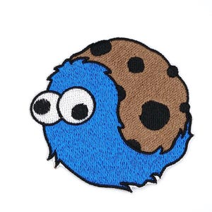 Könnte beinhalten: Ein gestickter Aufnäher, der ein rundes, flauschiges blaues Wesen mit zwei großen weißen Augen und schwarzen Pupillen zeigt, das teilweise von einem braunen Keks mit dunkelbraunen Flecken verdeckt wird. Das Design hat einen dicken schwarzen Rand.