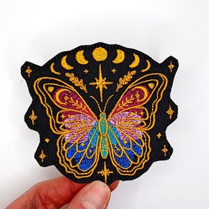 May include: Embroidered iron-on patch featuring a colorful butterfly with gold accents, surrounded by stars and crescent moons on a black background.