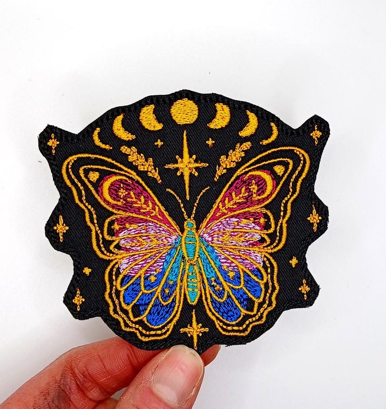 May include: Embroidered iron-on patch featuring a colorful butterfly with gold accents, surrounded by stars and crescent moons on a black background.
