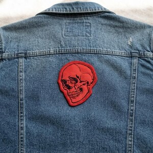 K&ouml;nnte beinhalten: Eine blaue Jeansjacke mit einem roten gestickten Aufn&auml;her auf der R&uuml;ckseite. Der Aufn&auml;her zeigt eine detaillierte schwarze Strichzeichnung eines menschlichen Sch&auml;dels auf rotem Grund mit einem roten Stickrand.