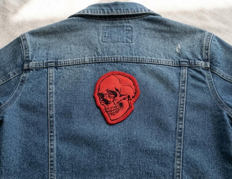 K&ouml;nnte beinhalten: Eine blaue Jeansjacke mit einem roten gestickten Aufn&auml;her auf der R&uuml;ckseite. Der Aufn&auml;her zeigt eine detaillierte schwarze Strichzeichnung eines menschlichen Sch&auml;dels auf rotem Grund mit einem roten Stickrand.