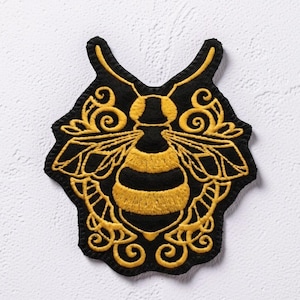 May include: A black and yellow embroidered patch featuring a central bee motif surrounded by intricate, symmetrical golden-yellow scrollwork and filigree patterns. The patch has a distinct, shaped border that follows the contours of the design.