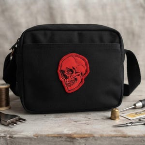 K&ouml;nnte beinhalten: Eine schwarze Umh&auml;ngetasche aus Segeltuch mit einem auff&auml;lligen, rot bestickten Totenkopf-Aufn&auml;her in der Mitte. Die Tasche hat eine rechteckige Form mit Rei&szlig;verschluss oben, einer Vordertasche und einem verstellbaren Schulterriemen.