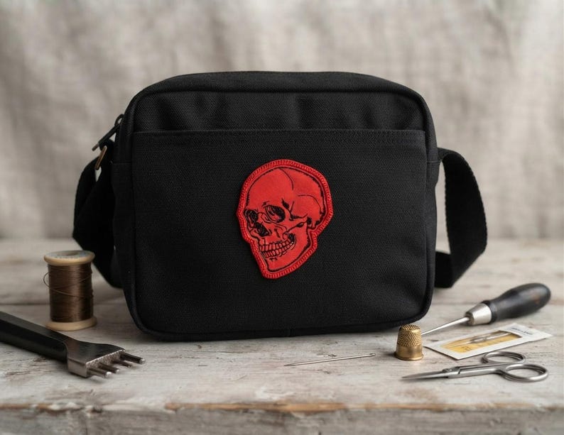 K&ouml;nnte beinhalten: Eine schwarze Umh&auml;ngetasche aus Segeltuch mit einem auff&auml;lligen, rot bestickten Totenkopf-Aufn&auml;her in der Mitte. Die Tasche hat eine rechteckige Form mit Rei&szlig;verschluss oben, einer Vordertasche und einem verstellbaren Schulterriemen.