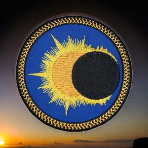 May include: A circular embroidered patch featuring a blue background with a central yellow sun partially eclipsed by a solid black circle. The patch is framed by a thick black border with a repeating gold-colored cross-stitch pattern.