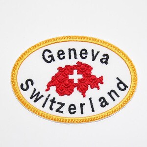 Könnte beinhalten: Ein weißer, ovaler gestickter Aufnäher mit goldener Umrandung. Der Aufnäher zeigt den Text "Genf Schweiz" und eine rote Umrandung der Schweiz mit einem weißen Kreuz in der Mitte.