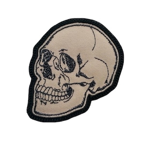 May include: Embroidered skull patch with a beige background and black border. The skull has detailed black stitching, including eye sockets, teeth, and facial features. The patch is designed for clothing or accessories.