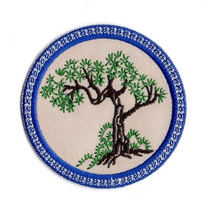 May include: Round embroidered patch with a tree design. The tree has green leaves and a brown trunk, set against a beige backdrop. It's encircled by a blue border with white stitching. Suitable for application on garments or accessories.
