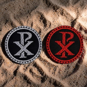 May include: Two circular embroidered patches featuring the Chi Rho symbol. One has a black background with a white Chi Rho and border. The other has a black background with a red Chi Rho and border. Both are displayed on a sandy surface.