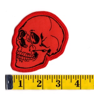 K&ouml;nnte beinhalten: Ein roter gestickter Aufn&auml;her in Form eines menschlichen Sch&auml;dels mit schwarzen Liniendetails. Der Aufn&auml;her liegt &uuml;ber einem gelben Ma&szlig;band, das zeigt, dass der Sch&auml;del etwa 9 cm breit und 10 cm hoch ist.