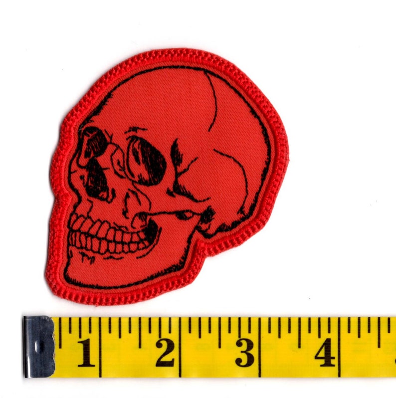 K&ouml;nnte beinhalten: Ein roter gestickter Aufn&auml;her in Form eines menschlichen Sch&auml;dels mit schwarzen Liniendetails. Der Aufn&auml;her liegt &uuml;ber einem gelben Ma&szlig;band, das zeigt, dass der Sch&auml;del etwa 9 cm breit und 10 cm hoch ist.