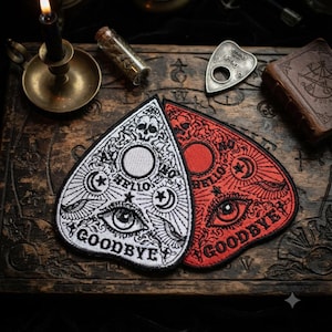 May include: Two embroidered patches shaped like planchette pointers, one in white with black thread and one in red with black thread. Both feature a central eye, crescent moons, stars, a skull, and the words HELLO and GOODBYE in bold lettering.