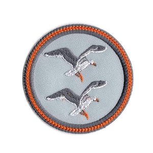 May include: A round, grey embroidered patch with an orange border. The patch features two white seagulls flying in a light blue sky.
