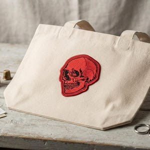 K&ouml;nnte beinhalten: Eine naturfarbene Canvas-Einkaufstasche mit einem mittig platzierten, roten, gestickten Totenkopf-Aufn&auml;her mit schwarzen Linien. Die Tasche hat zwei Tragegriffe und liegt neben einer roten Garnrolle, einem Fingerhut, Nadeln und einer Schere.