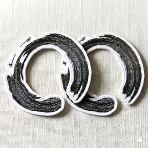 Könnte beinhalten: Zwei kreisförmige gestickte Aufnäher mit einem schwarzen Pinselstrich-Design im Tusche-Stil auf weißem Hintergrund. Jeder Aufnäher zeigt eine texturierte, handgezeichnete Optik mit variierender Strichstärke und einem weißen Rand.