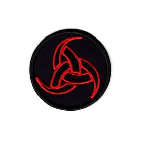 May include: A black and red embroidered patch with a circular design. The center of the patch features a red, three-pointed symbol with curved edges.