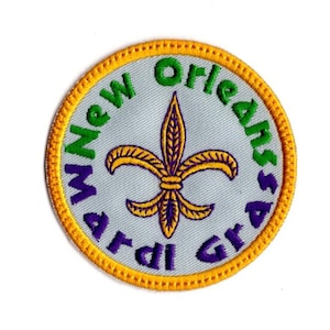 May include: Embroidered patch with a light gray background, featuring a gold fleur-de-lis and text that reads "New Orleans Mardi Gras" in green and purple. The patch has a gold border.