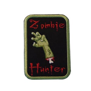 Op de afbeelding: Geborduurde rechthoekige patch met een zwarte achtergrond en een groene rand. De patch toont de woorden "Zombie Hunter" in rode letters en een groene zombiehand met een afgehakt polsgewricht.
