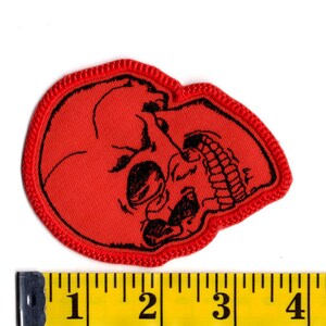 K&ouml;nnte beinhalten: Ein roter bestickter Aufn&auml;her mit einer schwarzen Strichzeichnung eines menschlichen Sch&auml;dels im Profil. Der Aufn&auml;her hat einen roten gestickten Rand und liegt &uuml;ber einem gelben Ma&szlig;band, das eine Breite von etwa 10 cm anzeigt.