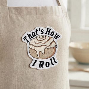 Könnte beinhalten: Eine beige Schürze mit einem gestickten Aufnäher einer Zimtschnecke mit weißem Zuckerguss. Der Text "That's How I Roll" ist in schwarzen Buchstaben über und unter der Zimtschnecken-Grafik auf den hellbeigen Stoff gestickt.