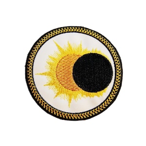 Könnte beinhalten: Ein runder gestickter Aufnäher mit goldener Umrandung und weißem Hintergrund. Der Aufnäher zeigt ein stilisiertes Bild einer Sonnenfinsternis mit einer gelben Sonne und einem schwarzen Mond.