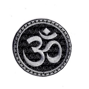 May include: A black and white embroidered patch with a silver Om symbol in the center. The background is a repeating pattern of small squares.