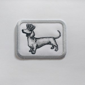 May include: A white embroidered patch featuring a black and white image of a dachshund wearing a crown. The patch has a silver border.