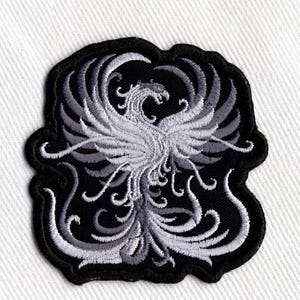 May include: Embroidered patch with a phoenix design. The patch has a black border and a detailed white and grey phoenix in the centre. The phoenix has outstretched wings and intricate feather details, suitable for jackets or bags.