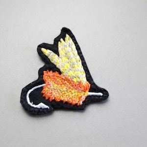 May include: An embroidered patch featuring a black silhouette of a cat with a long tail. The cat's body is adorned with a vibrant orange and yellow textured leaf pattern, while the ears and upper head area feature a pale yellow and white design.