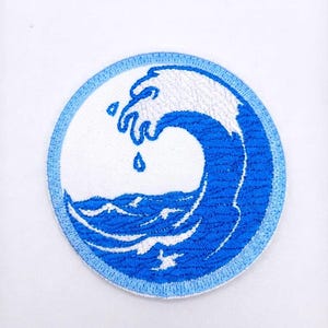 May include: A circular embroidered patch featuring a stylised blue and white ocean wave design. The patch has a light blue border, a white background, and a central graphic of a cresting wave with dark blue water and white foam details.