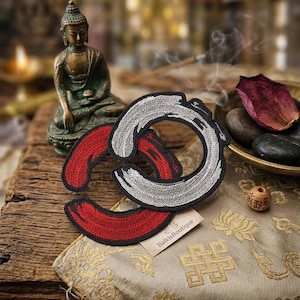May include: Embroidered patches in red and silver, with a black outline, on a decorative pillow with gold floral patterns. A small Buddha statue and a wooden bead with etched symbols are also visible.