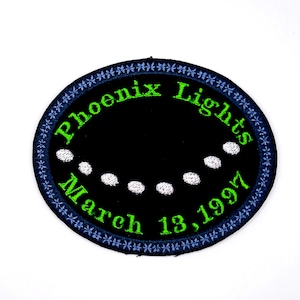 May include: Embroidered patch with a black oval center featuring the text "Phoenix Lights March 13, 1997" in green. The oval is surrounded by a blue border with a floral pattern and white dots.
