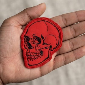 K&ouml;nnte beinhalten: Ein roter Stoffaufn&auml;her in Form eines menschlichen Sch&auml;dels. Der Aufn&auml;her verf&uuml;gt &uuml;ber eine detaillierte schwarze Stickkontur und Schattierungen, die die Augenh&ouml;hlen, die Nasenh&ouml;hle und die Z&auml;hne definieren. Die Kanten des sch&auml;delf&ouml;rmigen Aufn&auml;hers sind mit einem roten Rand versehen.