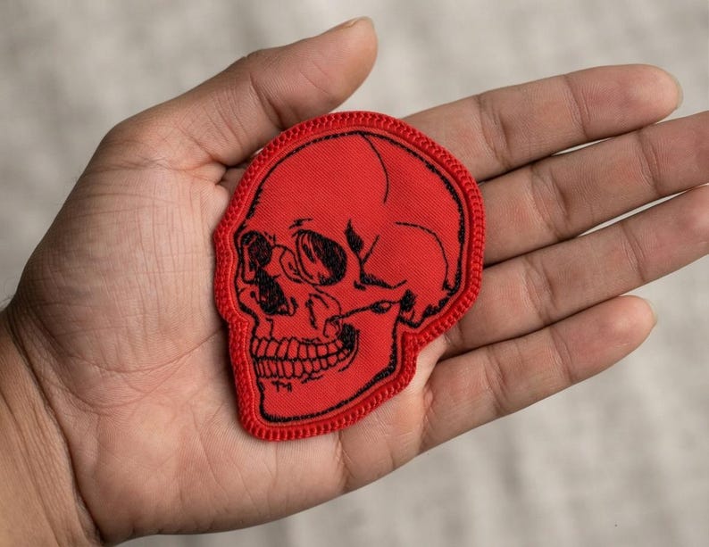 K&ouml;nnte beinhalten: Ein roter Stoffaufn&auml;her in Form eines menschlichen Sch&auml;dels. Der Aufn&auml;her verf&uuml;gt &uuml;ber eine detaillierte schwarze Stickkontur und Schattierungen, die die Augenh&ouml;hlen, die Nasenh&ouml;hle und die Z&auml;hne definieren. Die Kanten des sch&auml;delf&ouml;rmigen Aufn&auml;hers sind mit einem roten Rand versehen.