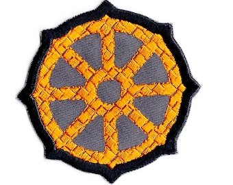 May include: A circular embroidered patch featuring a dark gray background with a bright orange eight-spoked wheel design. The patch has a thick, scalloped black border that frames the central orange and gray wheel pattern.