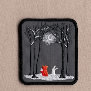 May include: Embroidered patch featuring a nighttime forest scene. A red fox and a white rabbit stand in a snowy clearing between black trees. A crescent moon shines above. The patch has a black border and is set against a neutral background.