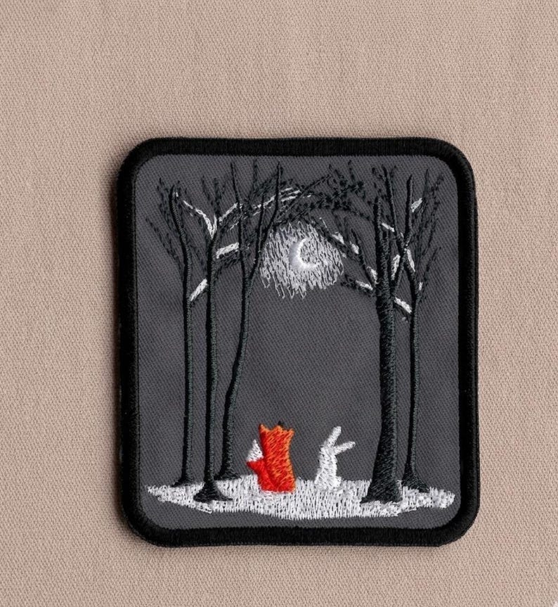 May include: Embroidered patch featuring a nighttime forest scene. A red fox and a white rabbit stand in a snowy clearing between black trees. A crescent moon shines above. The patch has a black border and is set against a neutral background.