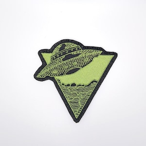 Könnte beinhalten: Gestickter Aufnäher mit einem grünen UFO über einer Landschaft, innerhalb eines schwarz umrandeten Dreiecks. Das UFO ist schwarz mit detaillierten Stickereien, der Hintergrund ist leuchtend grün. Geeignet für Kleidung oder Accessoires.