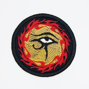 May include: Embroidered patch featuring the Eye of Horus symbol. The design includes a black border, a gold-colored eye, and red flame-like accents. The patch is circular and likely intended for application on clothing or accessories.