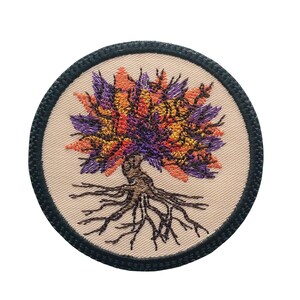 Könnte beinhalten: Runder, gestickter Aufnäher mit einem stilisierten Baum. Der Baum hat eine farbenfrohe Krone in Orange-, Gelb- und Lilatönen. Der Stamm und die Wurzeln sind braun, vor einem beigefarbenen Hintergrund und von einem schwarzen Rand umgeben. Ein dekorativer Artikel.
