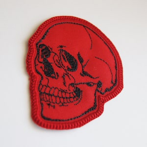 K&ouml;nnte beinhalten: Ein roter gestickter Aufn&auml;her in Form eines menschlichen Sch&auml;dels. Das Design zeigt schwarze Fadenstickereien, die die Augenh&ouml;hlen, die Nasenh&ouml;hle und die Z&auml;hne nachzeichnen, mit einem dicken roten Stickrand um die gesamte Sch&auml;delform.