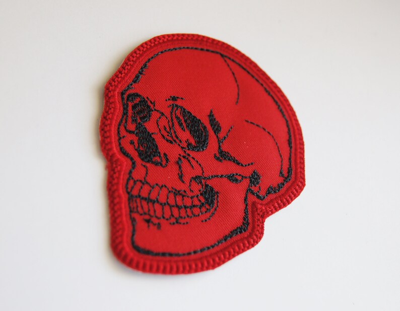 K&ouml;nnte beinhalten: Ein roter gestickter Aufn&auml;her in Form eines menschlichen Sch&auml;dels. Das Design zeigt schwarze Fadenstickereien, die die Augenh&ouml;hlen, die Nasenh&ouml;hle und die Z&auml;hne nachzeichnen, mit einem dicken roten Stickrand um die gesamte Sch&auml;delform.