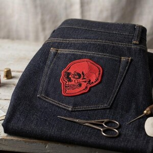 K&ouml;nnte beinhalten: Dunkelblaue Jeans aus Indigo-Denim mit einem roten, gestickten Totenkopf-Aufn&auml;her auf der Ges&auml;&szlig;tasche. Die Jeans ist von N&auml;hutensilien umgeben, darunter eine Spule mit rotem Garn, ein Fingerhut aus Metall, Nadeln, eine kleine Schere, eine Ahle und Schneiderkreide.