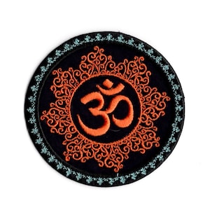 May include: A round embroidered patch with an orange Om symbol in the center. The Om symbol is surrounded by a decorative orange mandala design on a black background. The patch has a teal blue border.