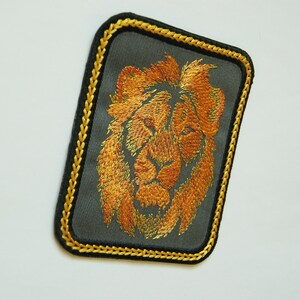 May include: A gold embroidered lion's head patch on a dark gray background with a gold stitched border.