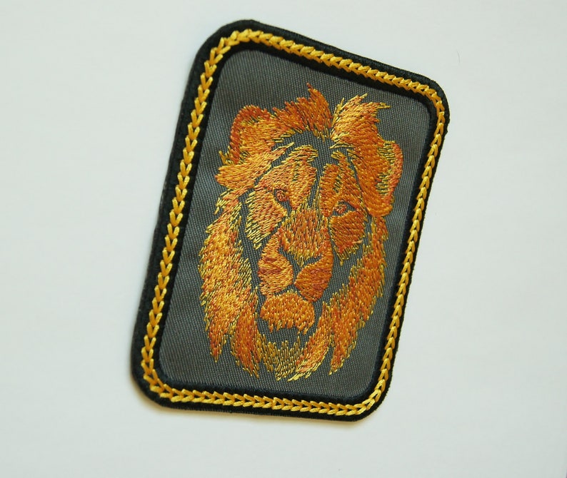 May include: A gold embroidered lion's head patch on a dark gray background with a gold stitched border.