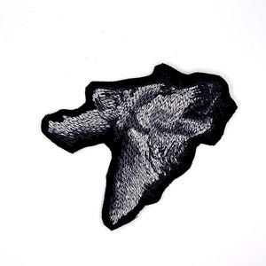 May include: A black and white embroidered patch in the shape of a wolf's head. The patch features detailed stitching to create a textured, realistic appearance. The wolf's head is set against a black border.