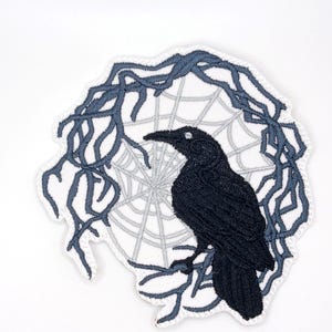 May include: An embroidered patch featuring a black raven perched on a branch, set against a light grey spider web design. The entire circular motif is framed by dark grey, tangled, leafless branches with a white stitched border edge.