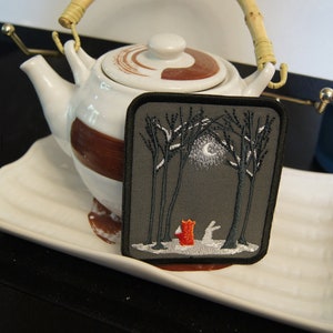 May include: Embroidered patch featuring a forest scene with a red fox and a white rabbit under a full moon. The patch is square with a black border and is set against a gray background. The patch is displayed on a white plate with a teapot in the background.