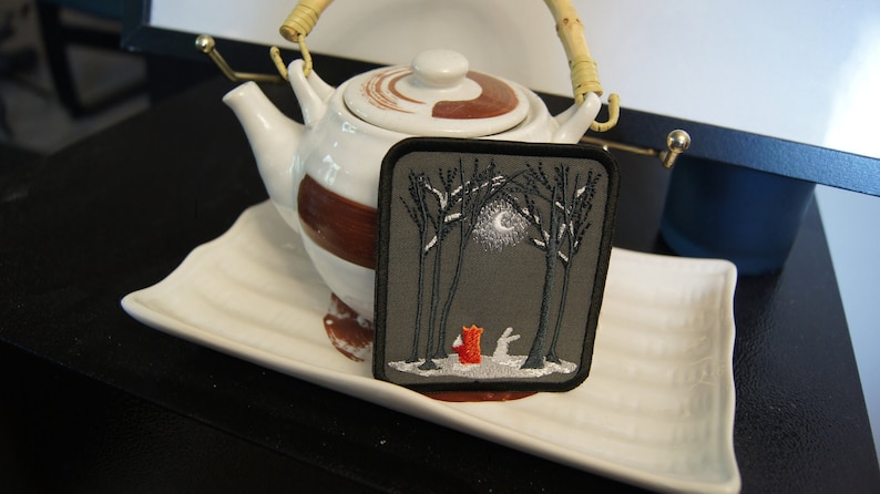 May include: Embroidered patch featuring a forest scene with a red fox and a white rabbit under a full moon. The patch is square with a black border and is set against a gray background. The patch is displayed on a white plate with a teapot in the background.
